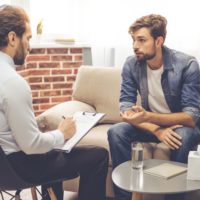 Handsome young man is sitting on couch and talking to the psychologist while doctor is making notes